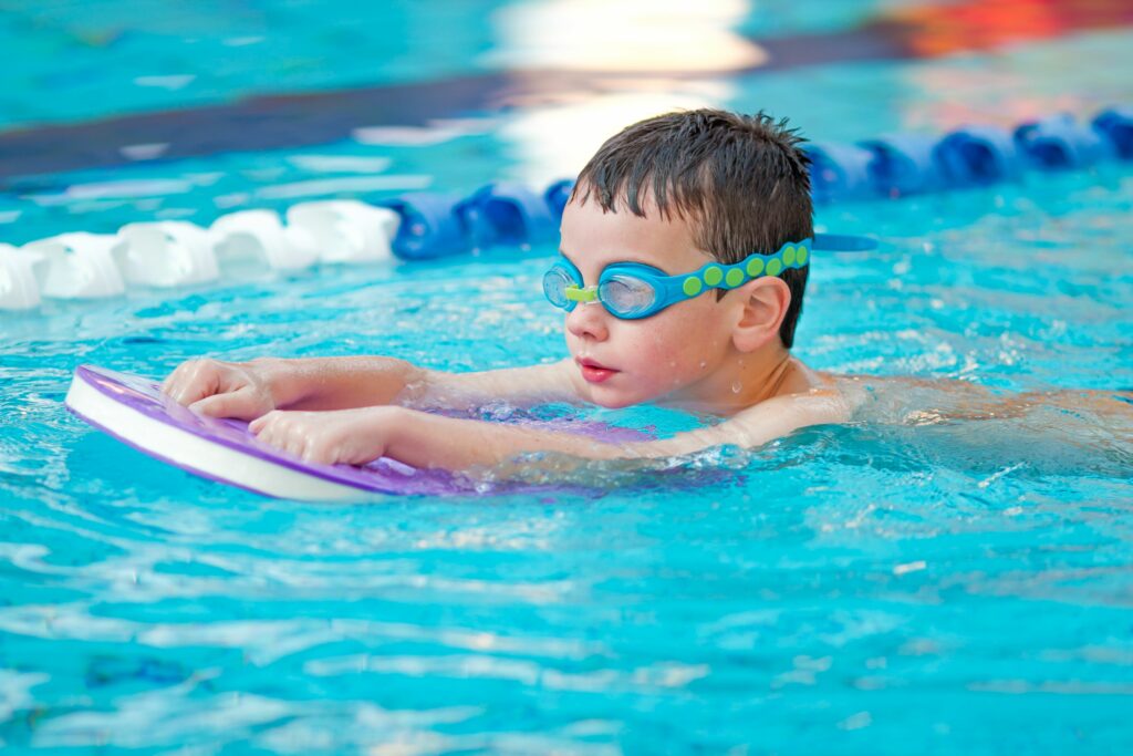 Pieni poika ui altaassa uimalaudan kanssa, uimalasit päässä.